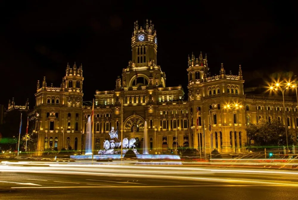 El Palacio de Cibeles iluminado de noche, con la fuente delante y las estelas de luz del tráfico