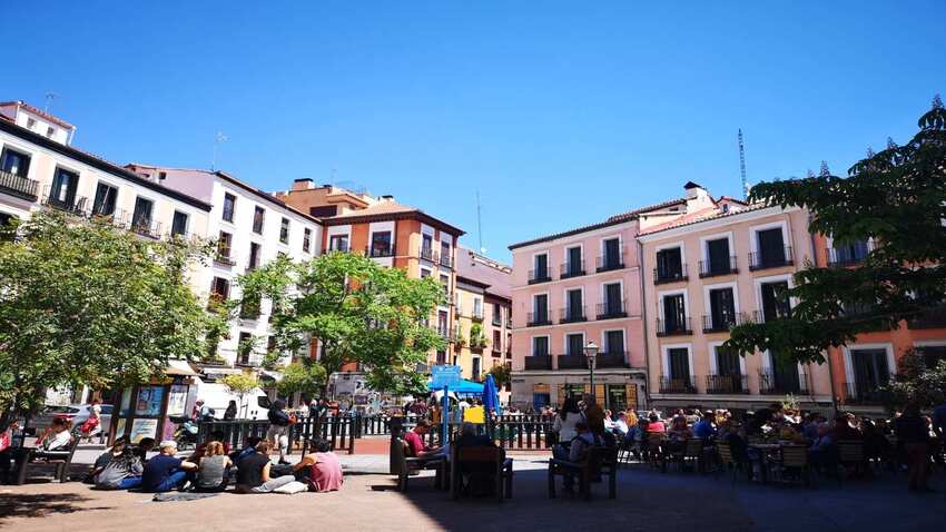 Gente sentada en los bancos y en las terrazas de una plaza de Malasaña rodeada de edificios de balcones